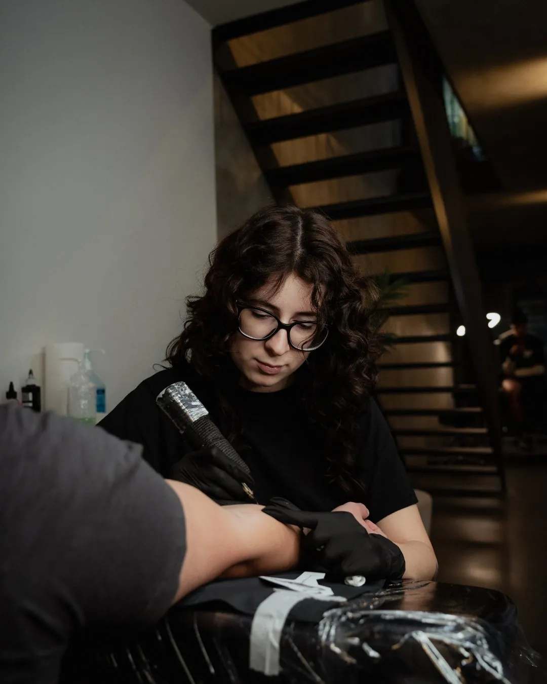 GON Berlin artist in glasses tattooing a client's forearm beneath the staircase